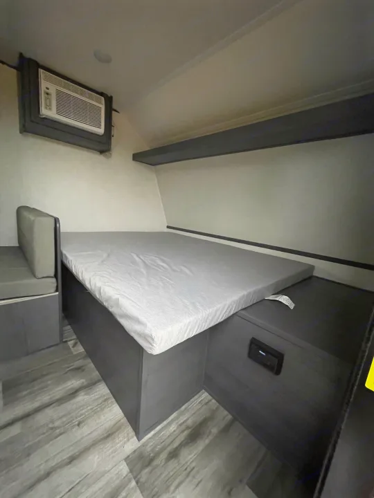Inside an RV rental: a compact sleeping area with a bed, bench, and wall AC unit for comfortable travel.. Dutchmen Aspen Trail 2023