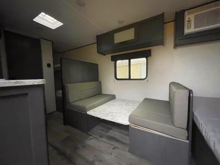 Inside this modern RV rental, a grey dinette with a marble-look table, overhead cabinets, and AC unit offer comfort.. Dutchmen Aspen Trail 2023