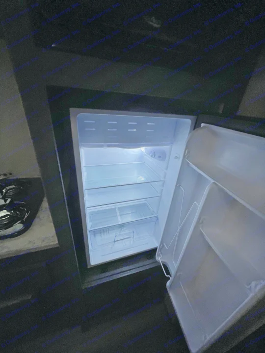 Inside an RV rental, an open, empty mini-fridge with clear shelves and a bright interior light.. Dutchmen Aspen Trail 2023