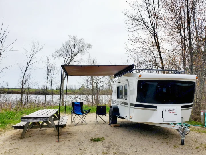 This RV rental is set up at a scenic lakeside campsite, complete with an awning, picnic table, and chairs.. Intech Luna Rover 2023
