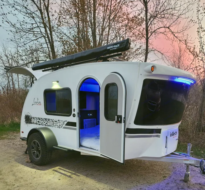 Experience an rv rental! This white inTech Luna Rover teardrop trailer features blue interior lights, an open door, and a rooftop awning under a pink sky.. Intech Luna Rover 2023