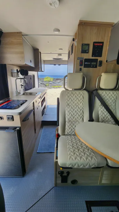 Modern rv rental interior: compact kitchen, seating with table, and control panels. A scenic ocean view is visible out the back.. Winnebago Solis 2023