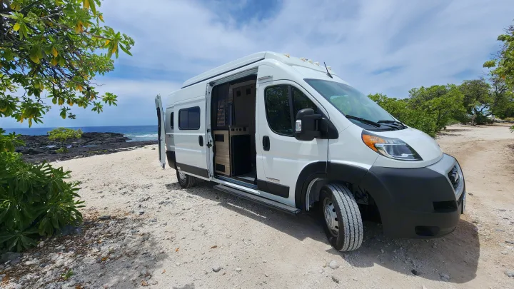 White RV rental camper van on a sandy, rocky beach. Ocean and volcanic shoreline in background. Perfect for nature adventures.. Winnebago Solis 2023