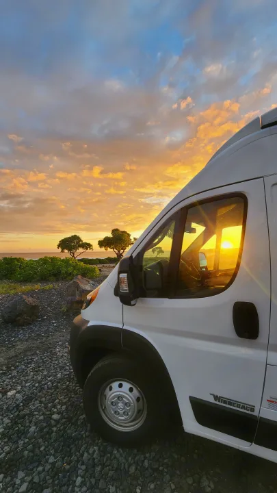 Golden sunset bathes an RV rental parked by the ocean. Ideal for a serene travel adventure on a rocky coast.. Winnebago Solis 2023