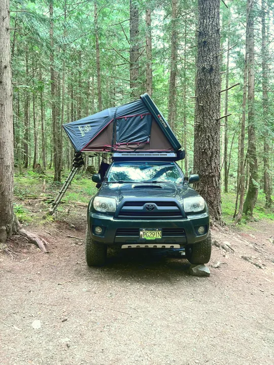 A dark green SUV with an iKamper rooftop tent and ladder is set up for forest camping. A unique rv rental option for wilderness adventures.. Toyota 4runner 2006