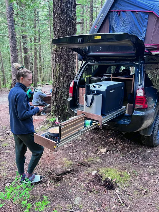 A person cooks on a slide-out stove from a custom SUV, equipped with a rooftop tent. Great for an rv rental outdoor adventure.. Toyota 4runner 2006