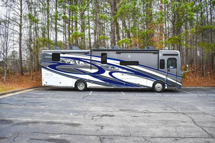 A sleek gray, white, and blue rv rental motorhome is parked in a lot with "NO PARKING" text, surrounded by autumn trees.. Holiday Rambler Endeavor 2022