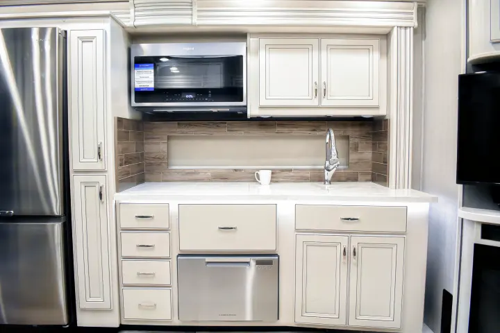 Modern RV rental kitchen: stainless fridge, microwave, dishwasher, white cabinets & counter, with a tiled backsplash.. Holiday Rambler Endeavor 2022