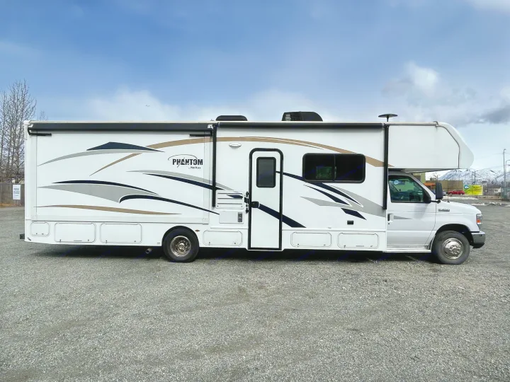 Full side view of a white Class C Phantom by NeXus RV rental with slide-out extended, parked on gravel.. Nexus Phantom 2018