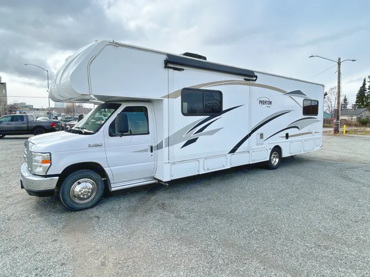 Side view of a white Phantom Nexus Class C RV, ideal for RV rental, parked on gravel under a cloudy sky.. Nexus Phantom 2018