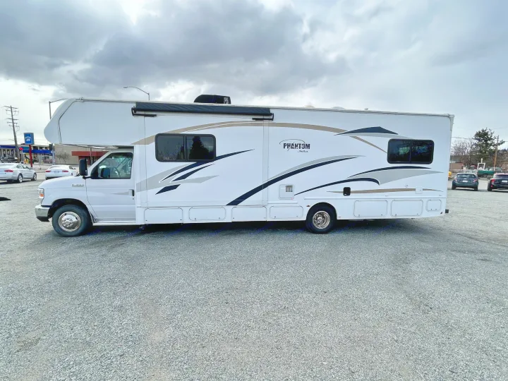 A white Class C RV rental with tan and black graphics is parked on a gravel lot under a cloudy sky, ready for adventure.. Nexus Phantom 2018