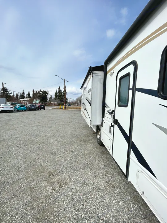 White RV rental with an extended slide-out parked on a gravel lot. Cars and houses in the background.. Nexus Phantom 2018