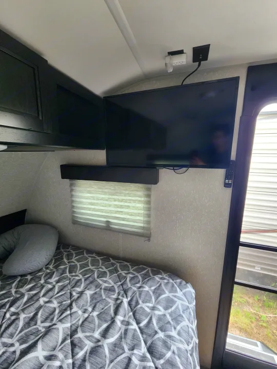 An RV rental bedroom features a wall-mounted TV, comfy bed with geometric comforter, and window with blinds.. Forest River Cherokee Wolf Pup Black Label 2022