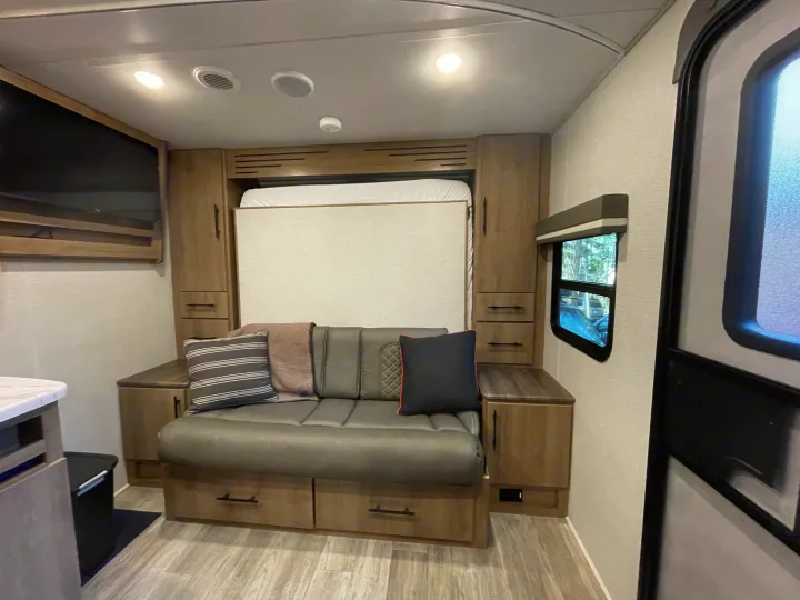 RV rental living area with a gray couch, hidden murphy bed, and light wood cabinets, plus a window.. Grand Design Imagine XLS 2023