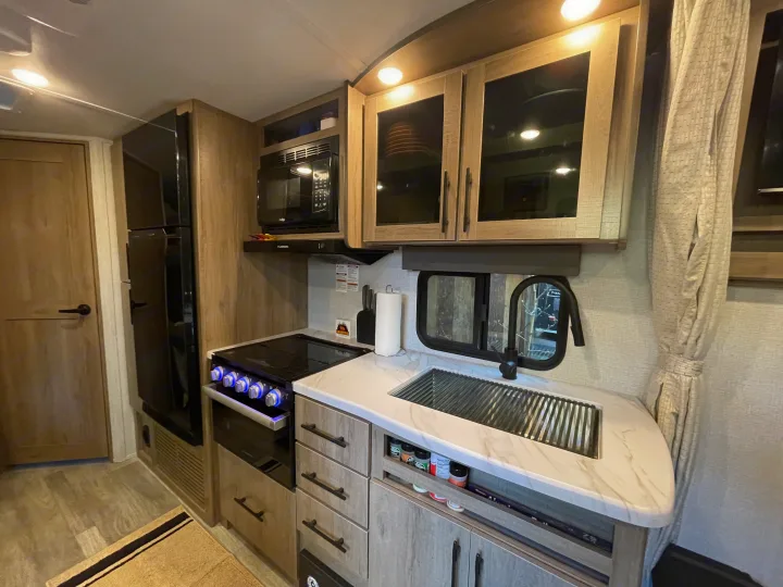 Experience comfort in this RV rental kitchen, featuring a modern stove, oven, microwave, large fridge, and a functional sink area.. Grand Design Imagine XLS 2023