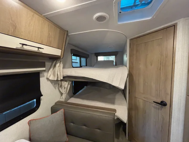 Inside an RV rental: two neatly made bunk beds, a couch in the foreground, wooden cabinets, and a light-colored door.. Grand Design Imagine XLS 2023