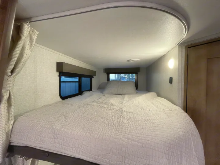 Relaxing RV rental bedroom: a large, neatly made bed, bright windows, and cozy interior lighting offer comfort on the road.. Grand Design Imagine XLS 2023