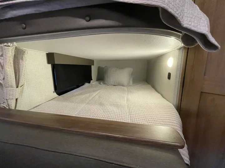 View of a compact sleeping area in an RV rental. Shows a neat bed, window with blind, and side light.. Grand Design Imagine XLS 2023