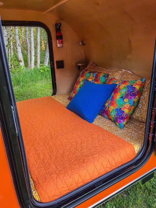 Cozy RV rental interior: Orange bed, colorful pillows, and a scenic forest view out the open door.. Aero Build Burnside 2023