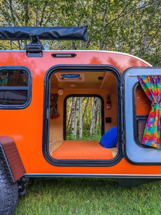 Orange teardrop RV rental in nature with open door revealing cozy bed & forest view. Ready for adventure.. Aero Build Burnside 2023