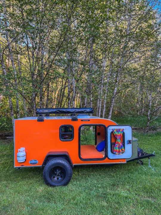 Vibrant orange teardrop camper, a perfect RV rental option, nestled in a tranquil forest. Explore nature!. Aero Build Burnside 2023
