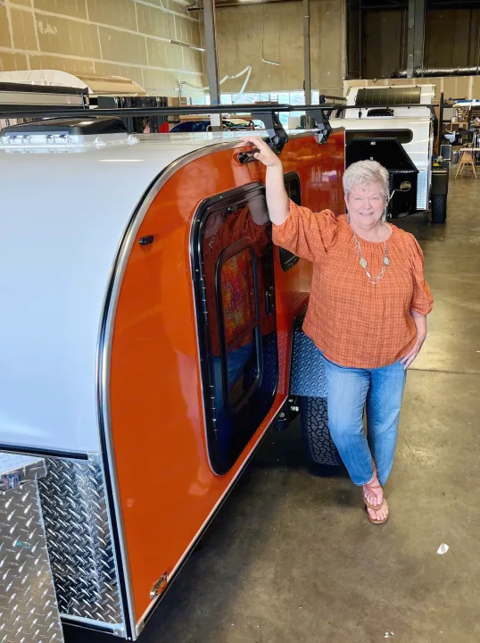 A smiling woman showcases a vibrant orange & white teardrop trailer, ideal for rv rental adventures. See this unique camper!. Aero Build Burnside 2023