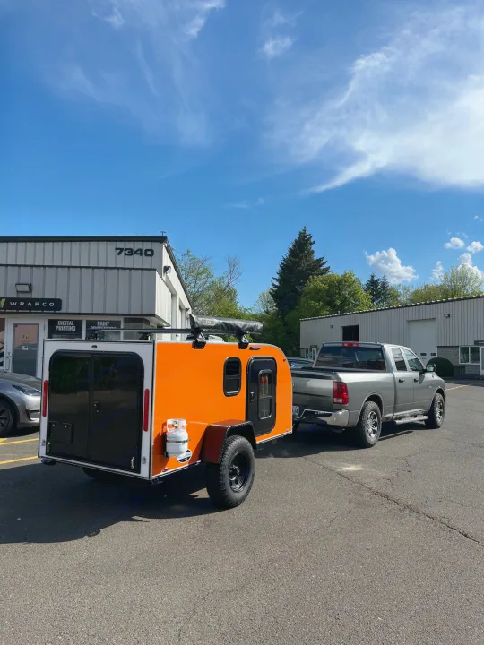 Bright orange teardrop RV rental hooked to a grey pickup truck in a sunny lot.. Aero Build Burnside 2023
