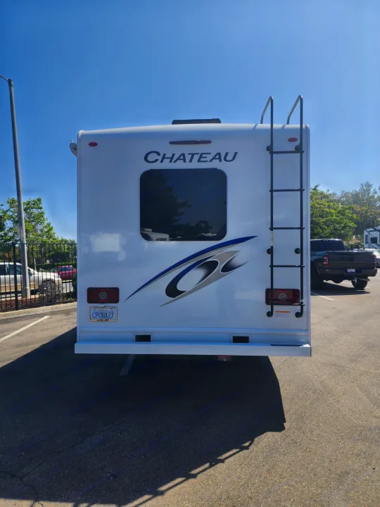 A white Chateau RV rental, seen from the rear with a ladder and window, is parked outdoors under a blue sky.. Thor Motor Coach Chateau 2023