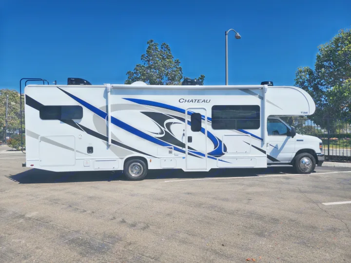 Bright white rv rental, a modern motorhome with dynamic blue and black graphics, parked on asphalt under a clear sky.. Thor Motor Coach Chateau 2023