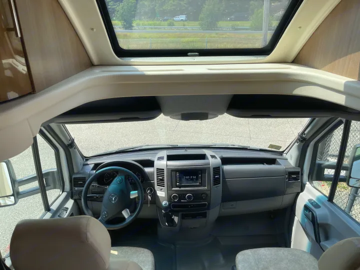 View from inside an RV rental showing the driver's cockpit with a Mercedes-Benz steering wheel, dashboard controls, and a panoramic overhead window. Road visible ahead.. Itasca Navion 2015