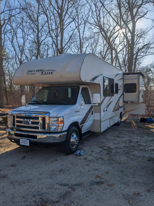 Explore the outdoors with this Class C RV rental. A white Freedom Elite camper is parked on a sunny dirt campsite among bare trees.. Thor Freedom Elite 2017