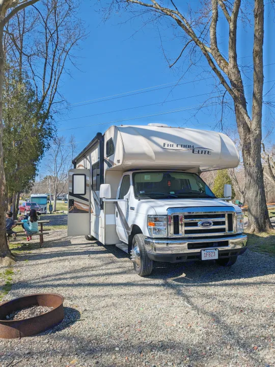 Perfect camping day! This Freedom Elite RV rental is set up on a gravel lot with a picnic table and fire pit. People relax nearby.. Thor Freedom Elite 2017