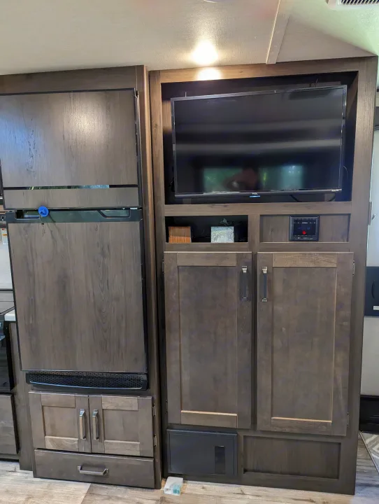 This RV rental features a dark wood-paneled refrigerator, a wall-mounted TV, and an entertainment center with cabinets.. Grand Design Transcend Xplor 2022