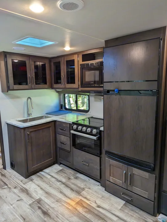 This RV rental kitchen features dark wood cabinets, a stainless sink, microwave, range, and full-size fridge.. Grand Design Transcend Xplor 2022