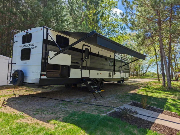A large white and grey travel trailer, ideal for an rv rental, is parked at a sunny woodland campsite with its awning out and steps extended.. Grand Design Transcend Xplor 2022