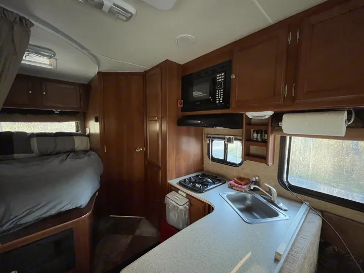 Explore this RV rental interior: a compact kitchen with stove, sink, and microwave, next to a cozy bed.. Coachmen Leprechaun 2014