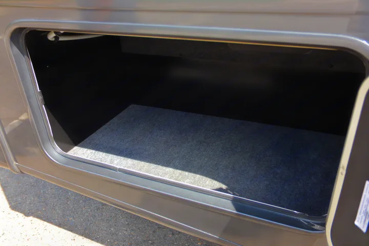 Open, empty exterior storage compartment of an RV rental, featuring a gray carpeted floor, perfect for packing your adventure gear.. Coachmen Encore 2024