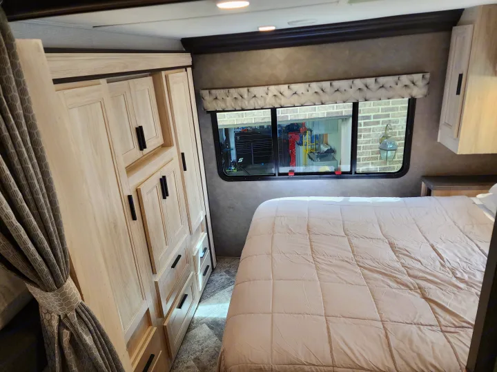 RV rental bedroom interior with light wood cabinets, a comfy bed, and window showing a brick wall outside. Ideal for travel.. Forest River Sunseeker LE 2021