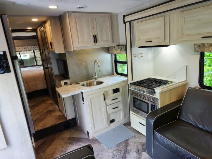 Bright rv rental interior showcasing a compact kitchen with stove, microwave, sink, adjacent sofa, and a bedroom beyond.. Forest River Sunseeker LE 2021