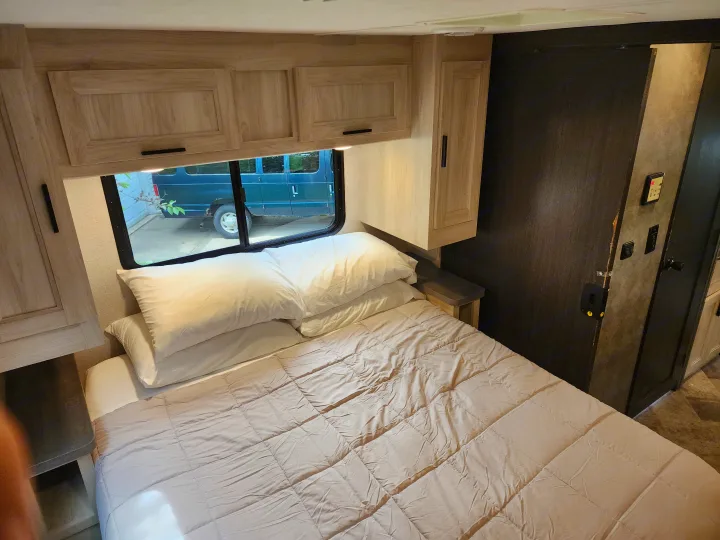 RV rental bedroom featuring a comfortable bed, overhead light wood cabinets, and a window revealing an outdoor van.. Forest River Sunseeker LE 2021