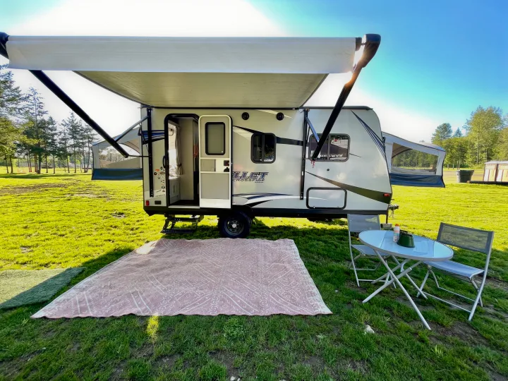 Perfect rv rental campsite: Travel trailer with open door, extended awning, outdoor rug, and table/chairs on lush green grass.. Keystone RV Bullet Crossfire 2019