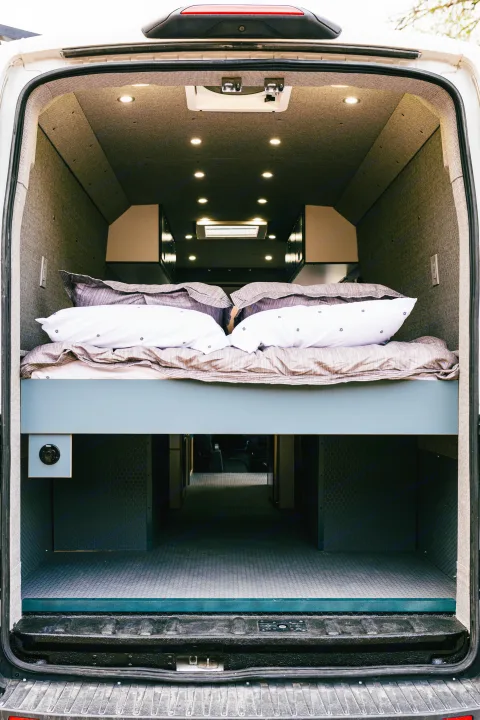 Rear view inside a modern RV rental, featuring a raised bed with pillows and sheets, and a large storage area below.. Ford Transit 250 RWD 2018