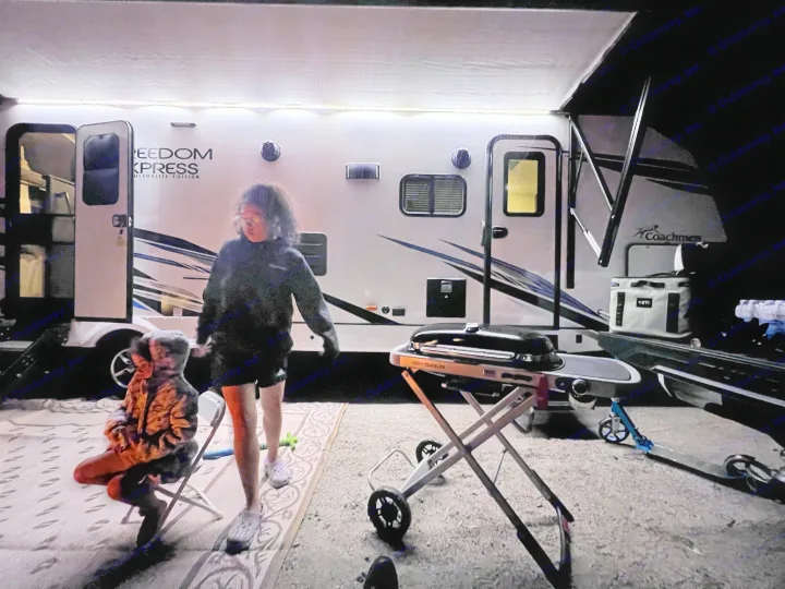 Nighttime at an RV rental: a white camper with a lit awning, people, a grill, and a cooler setup for outdoor comfort.. Coachmen freedom express 287bhds 2022