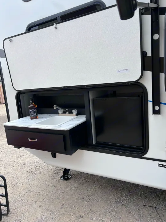 RV rental outdoor kitchen setup, revealing a pull-out sink, faucet, drawer, and mini-fridge. A whiskey bottle sits on the counter.. Coachmen freedom express 287bhds 2022