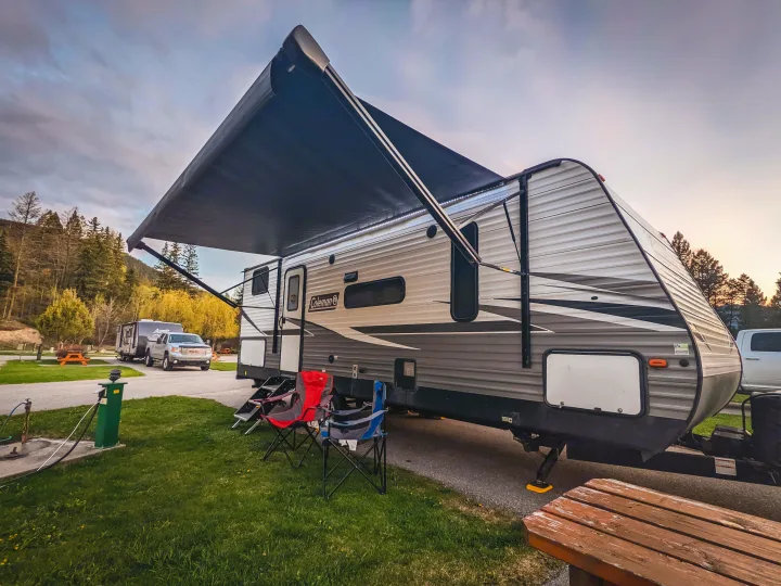Relaxing RV rental campsite: A travel trailer with an extended awning, chairs, and picnic table at dusk in a scenic campground.. Coleman Lantern 2021