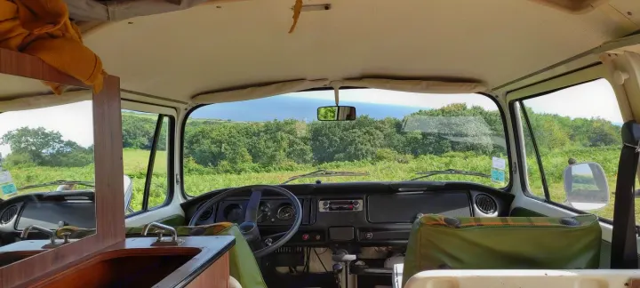 An inviting RV rental: Inside a vintage camper van, showing the dashboard and scenic outdoor views over green land and ocean.. Volkswagen T2 Westfalia 1976