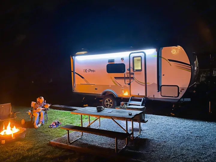 Night scene: Person and dog relax by a warm campfire beside a bright rv rental travel trailer and picnic table.. Forest River Flagstaff E-Pro 2023