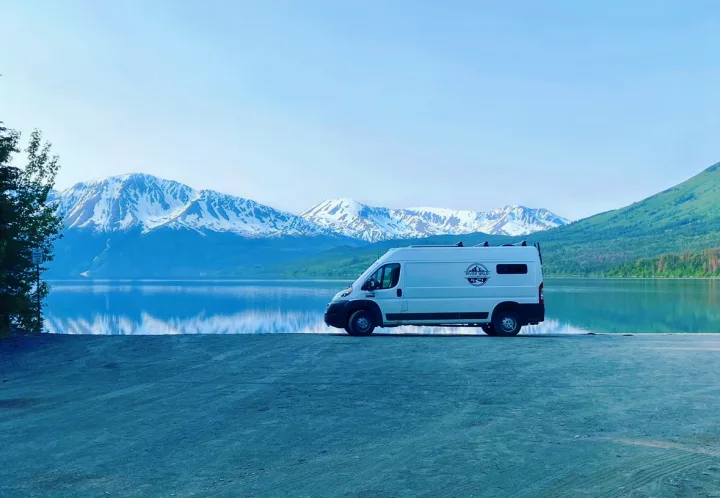 A white camper van, an RV rental, is parked by a calm lake that mirrors snow-capped mountains.. Dodge Promaster 2500 (Class B) 2022