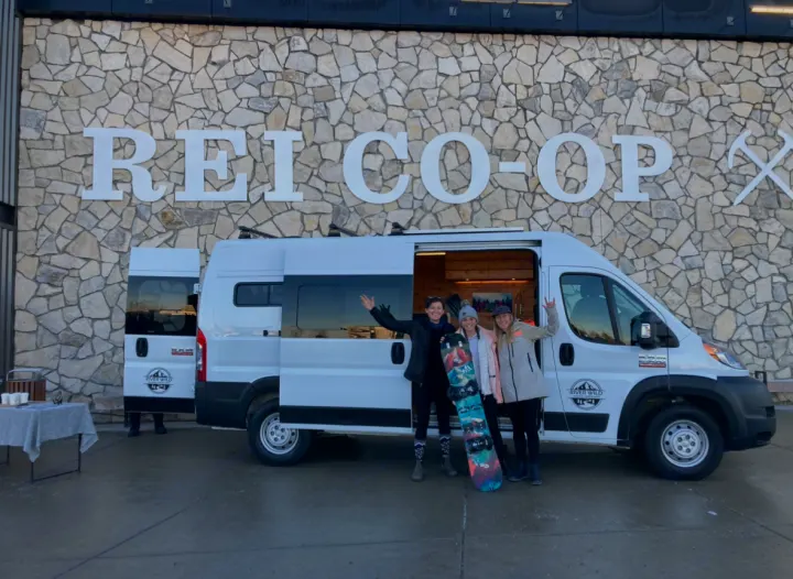 Three women with a snowboard and River Wild RV rental camper van at REI CO-OP, ready for adventure.. Dodge Promaster 2500 (Class B) 2022