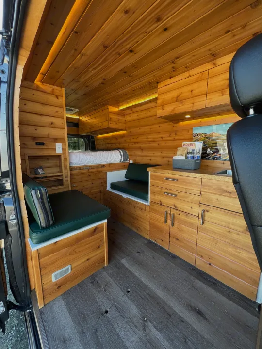 Bright RV rental interior features warm wood paneling, a comfy bed, kitchenette, and green seating in a modern camper van.. Dodge Promaster 2500 (Class B) 2022